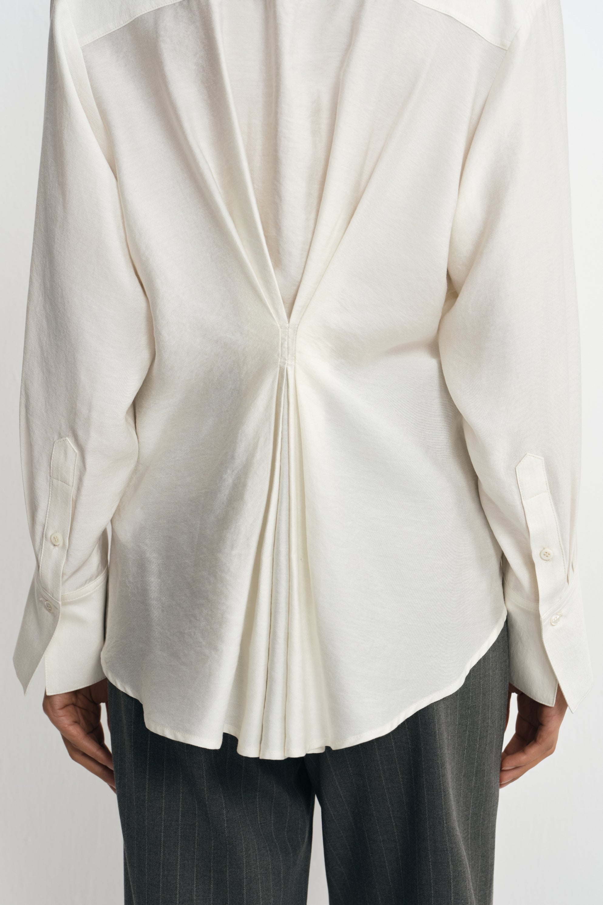 Close-up of a winter-white blouse on a model from behind, showing pleats drawn together at the waist and fanning out toward the curved hem.