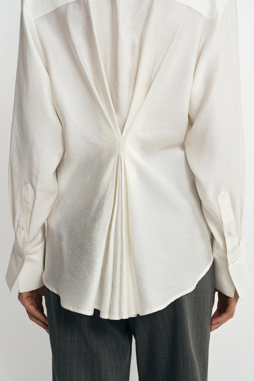 Close-up of a winter-white blouse on a model from behind, showing pleats drawn together at the waist and fanning out toward the curved hem.