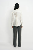 Back view of a model wearing a winter-white blouse with pleats gathered at the center waist, paired with charcoal pinstriped trousers.