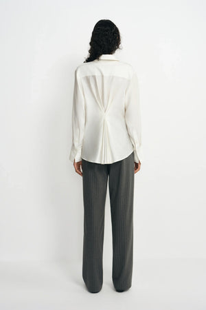 Back view of a model wearing a winter-white blouse with pleats gathered at the center waist, paired with charcoal pinstriped trousers.