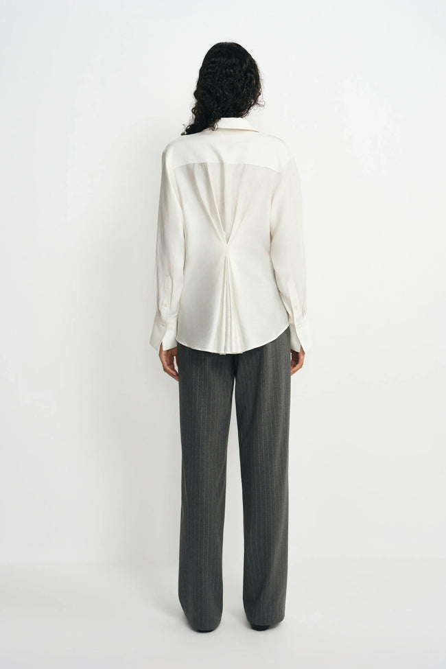 Back view of a model wearing a winter-white blouse with pleats gathered at the center waist, paired with charcoal pinstriped trousers.