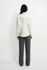 Back view of a model wearing a winter-white blouse with pleats gathered at the center waist, paired with charcoal pinstriped trousers.