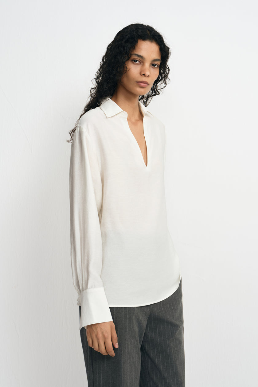 Angled close-up of a model wearing a winter-white blouse, showing the pointed collar, open neckline and gathered sleeve above a wide cuff.