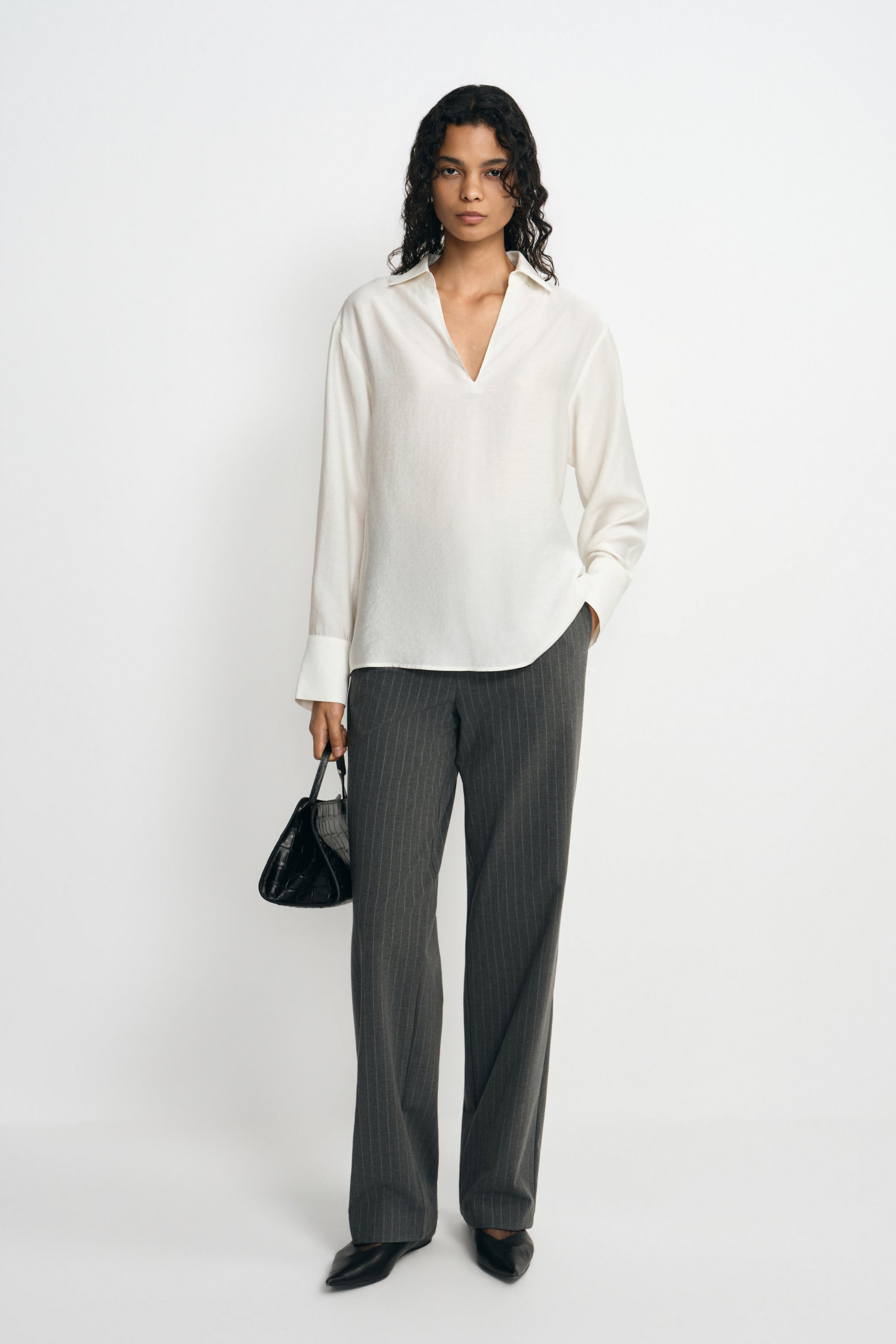 A model facing forward in a winter-white blouse with an open collar and curved hem, paired with charcoal pinstriped trousers, black shoes and a black bag.