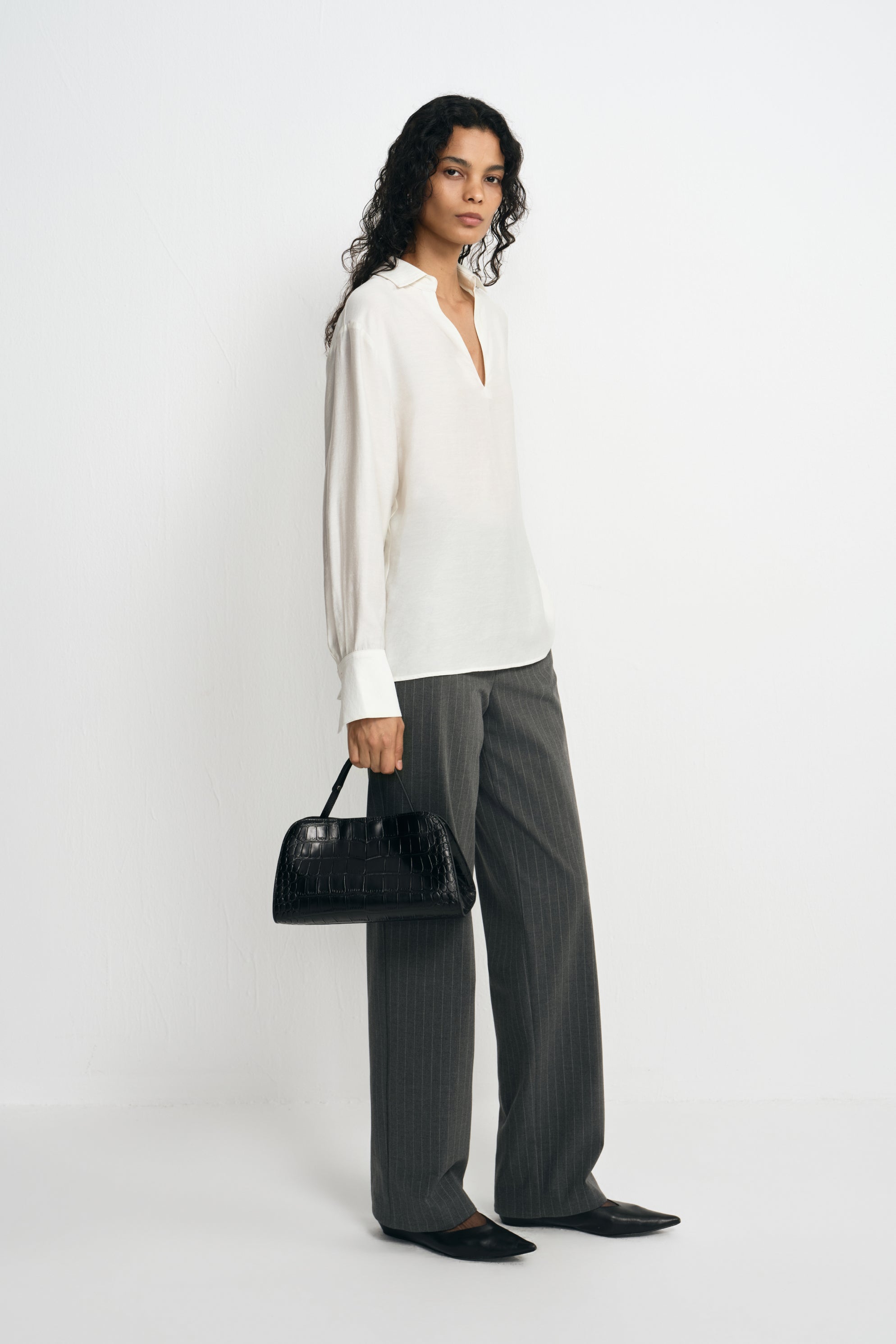 A model standing at an angle in a winter-white long-sleeve blouse with broad cuffs, styled with charcoal striped trousers, pointed shoes and a black handbag.