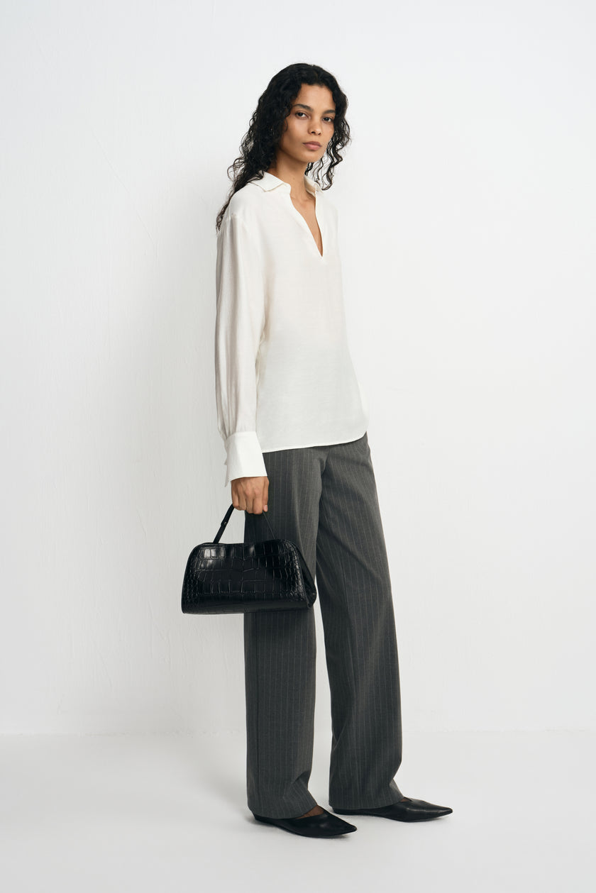 A model standing at an angle in a winter-white long-sleeve blouse with broad cuffs, styled with charcoal striped trousers, pointed shoes and a black handbag.