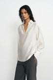 A model wearing a winter-white blouse with an open pointed collar, long sleeves and broad cuffs, worn untucked over charcoal pinstriped trousers.