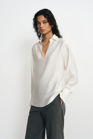 A model wearing a winter-white blouse with an open pointed collar, long sleeves and broad cuffs, worn untucked over charcoal pinstriped trousers.
