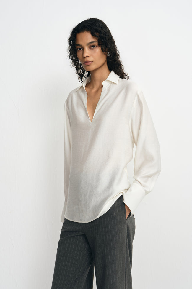 A model wearing a winter-white blouse with an open pointed collar, long sleeves and broad cuffs, worn untucked over charcoal pinstriped trousers.