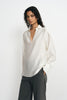 A model wearing a winter-white blouse with an open pointed collar, long sleeves and broad cuffs, worn untucked over charcoal pinstriped trousers.