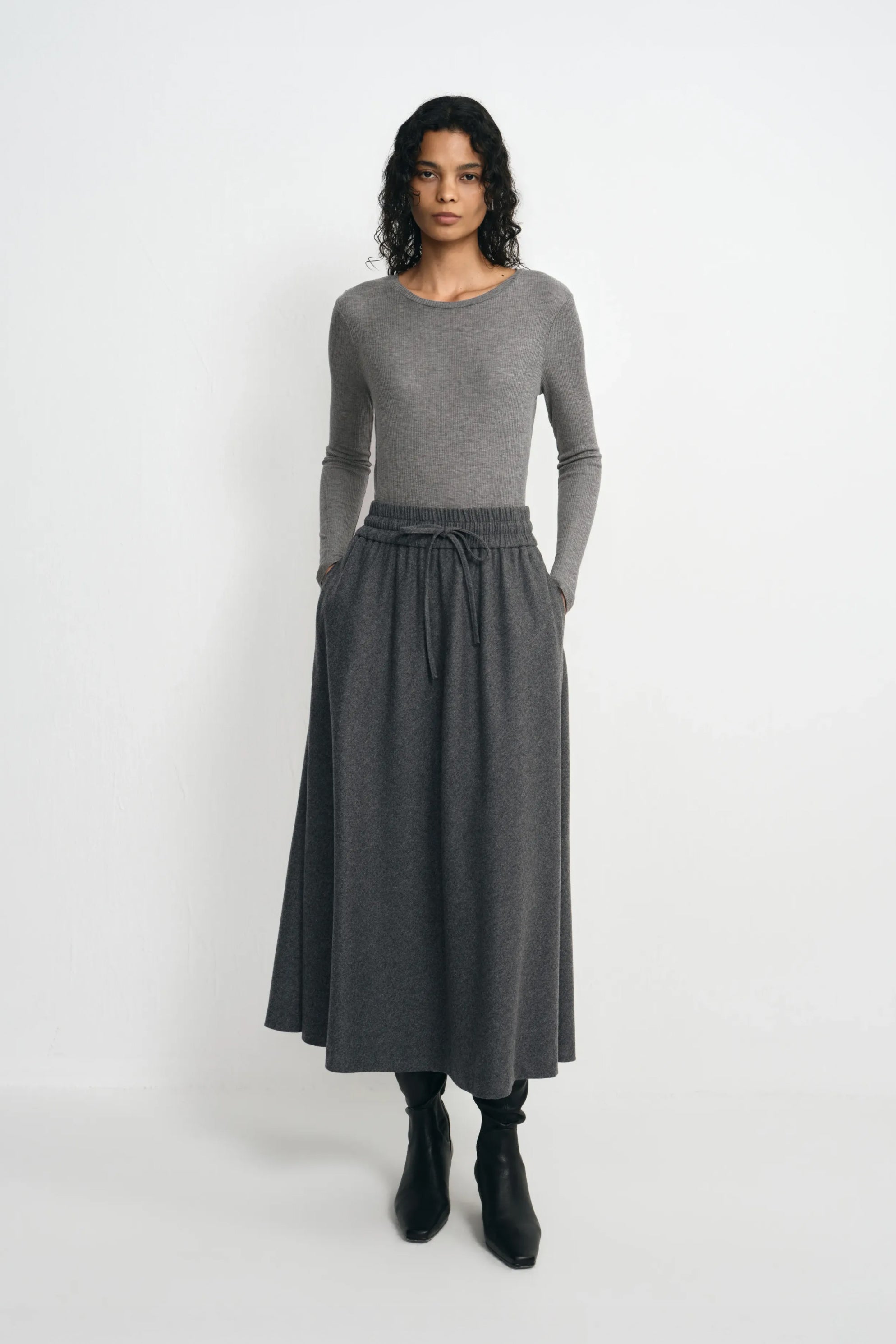 A model facing forward in a medium-grey ribbed top, tucked into a charcoal drawstring midi skirt and paired with black heeled boots.