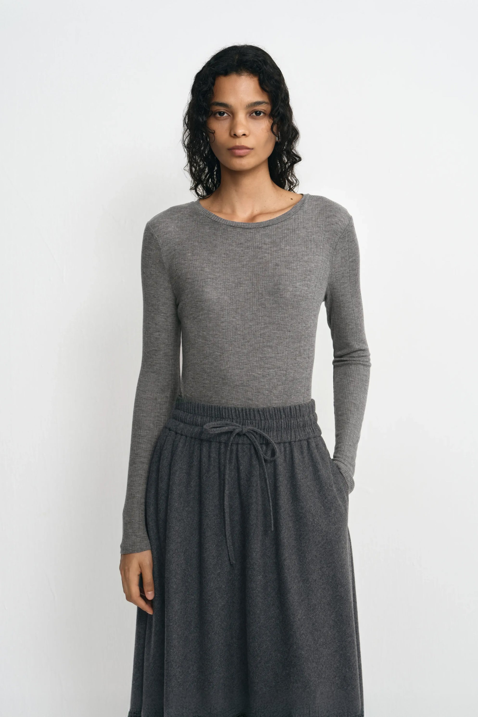 Close-up of a model wearing a medium-grey lyocell-wool top, showing fine ribbing, a narrow crewneck edge and long sleeves above a charcoal skirt.