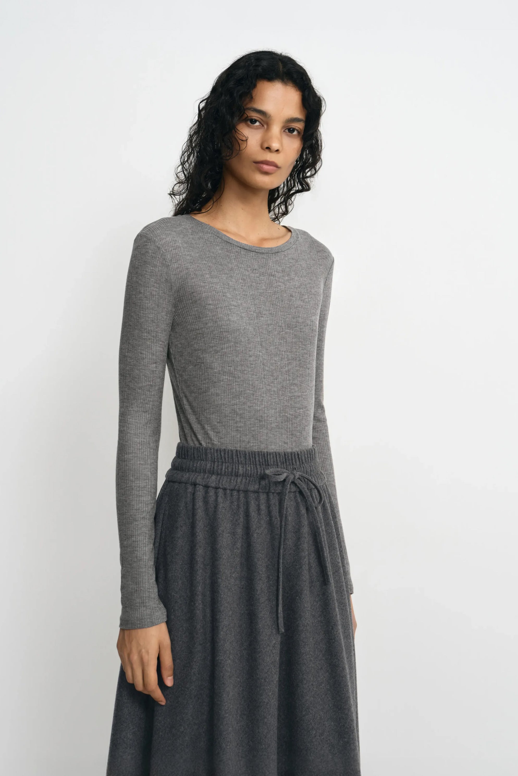 Three-quarter close-up of a model in a medium-grey ribbed top, showing the crew neck and narrow sleeves, with the hem tucked into a charcoal skirt.