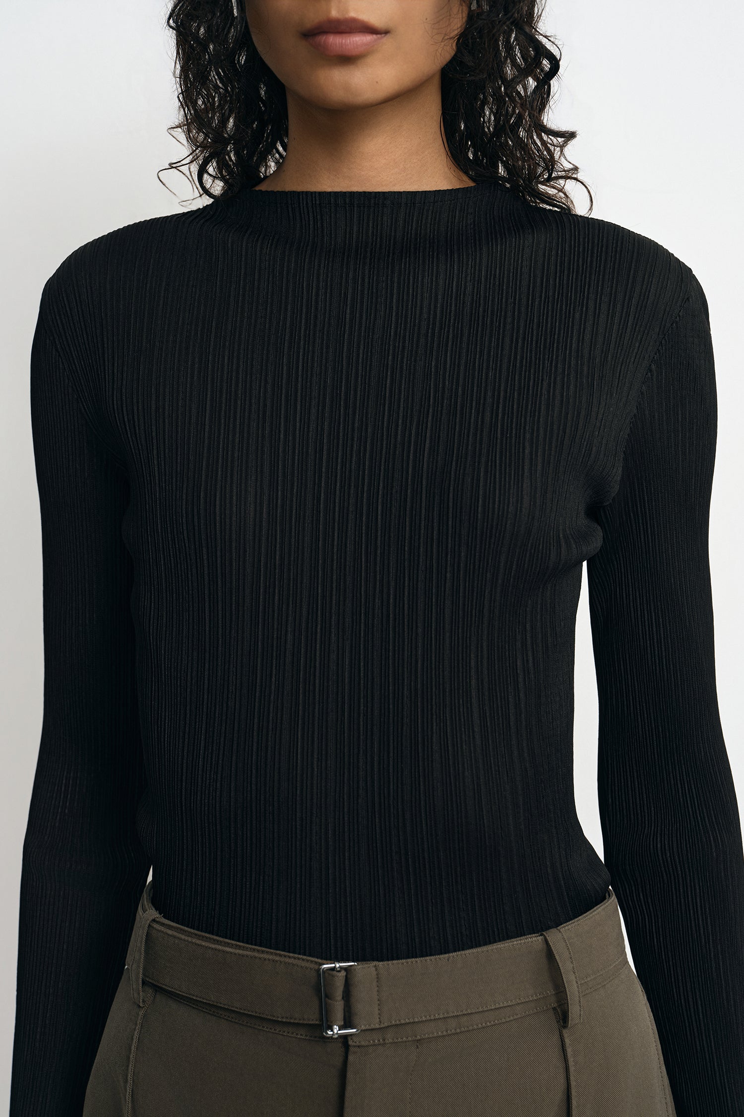 Close-up of a black pleated turtleneck top showing the ribbed texture at the neckline, paired with olive trousers with a buckled waist.
