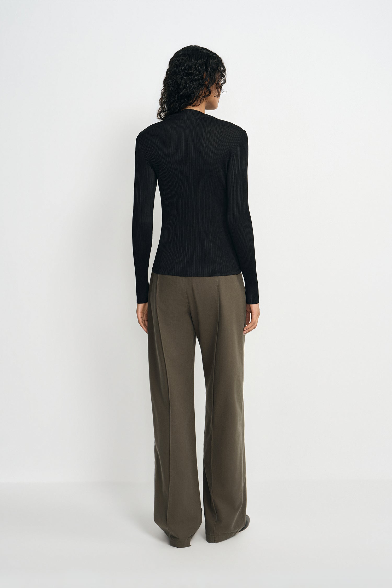 A model wearing a black pleated turtleneck top, pairing olive wide-leg trousers, back view.