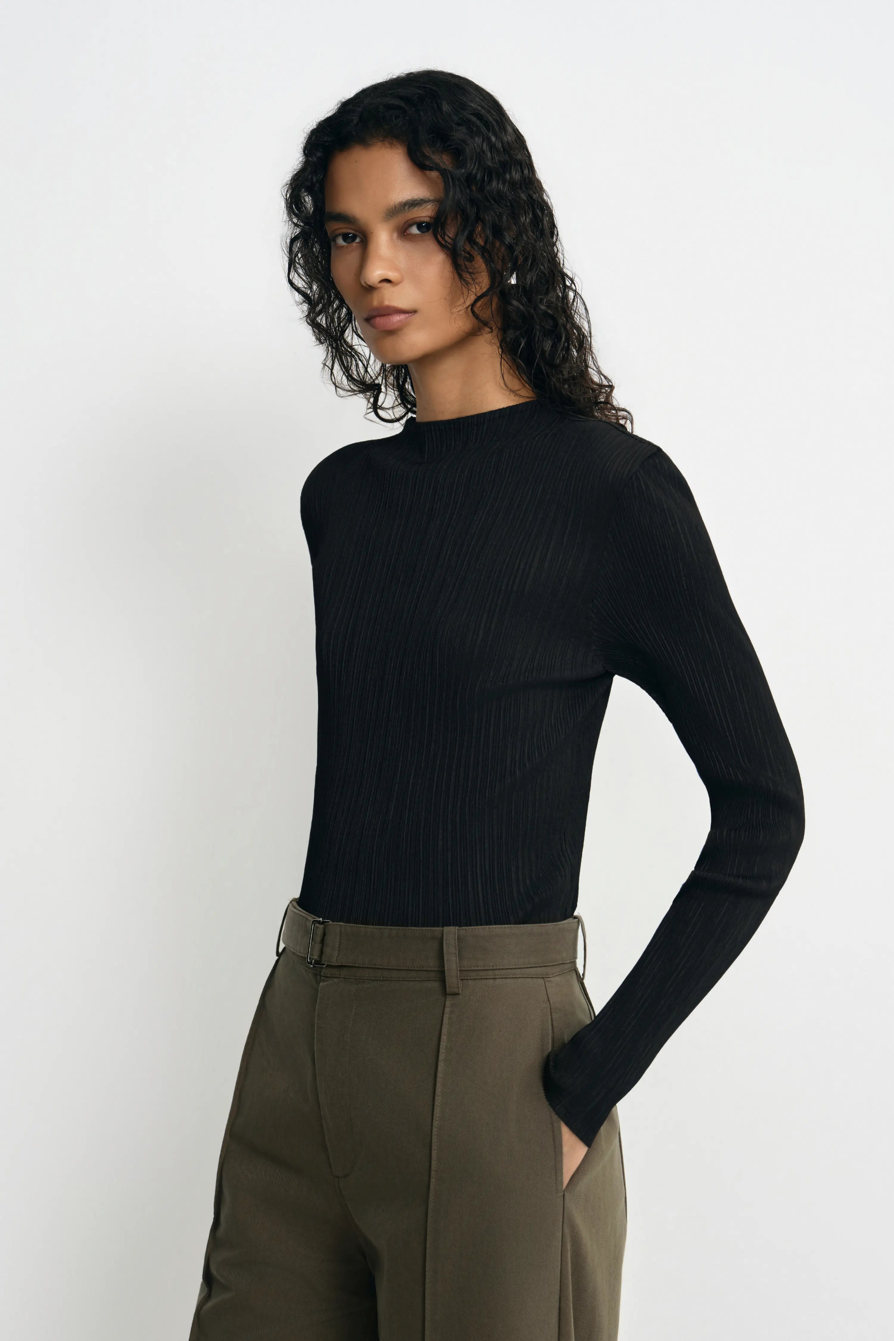A model wearing a black ribbed long-sleeve mock neck top and olive green trousers, standing against a plain light gray background.