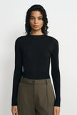 A woman modeling a black long-sleeve ribbed knit top with a boat neckline, paired with olive green trousers, against a plain light gray background.