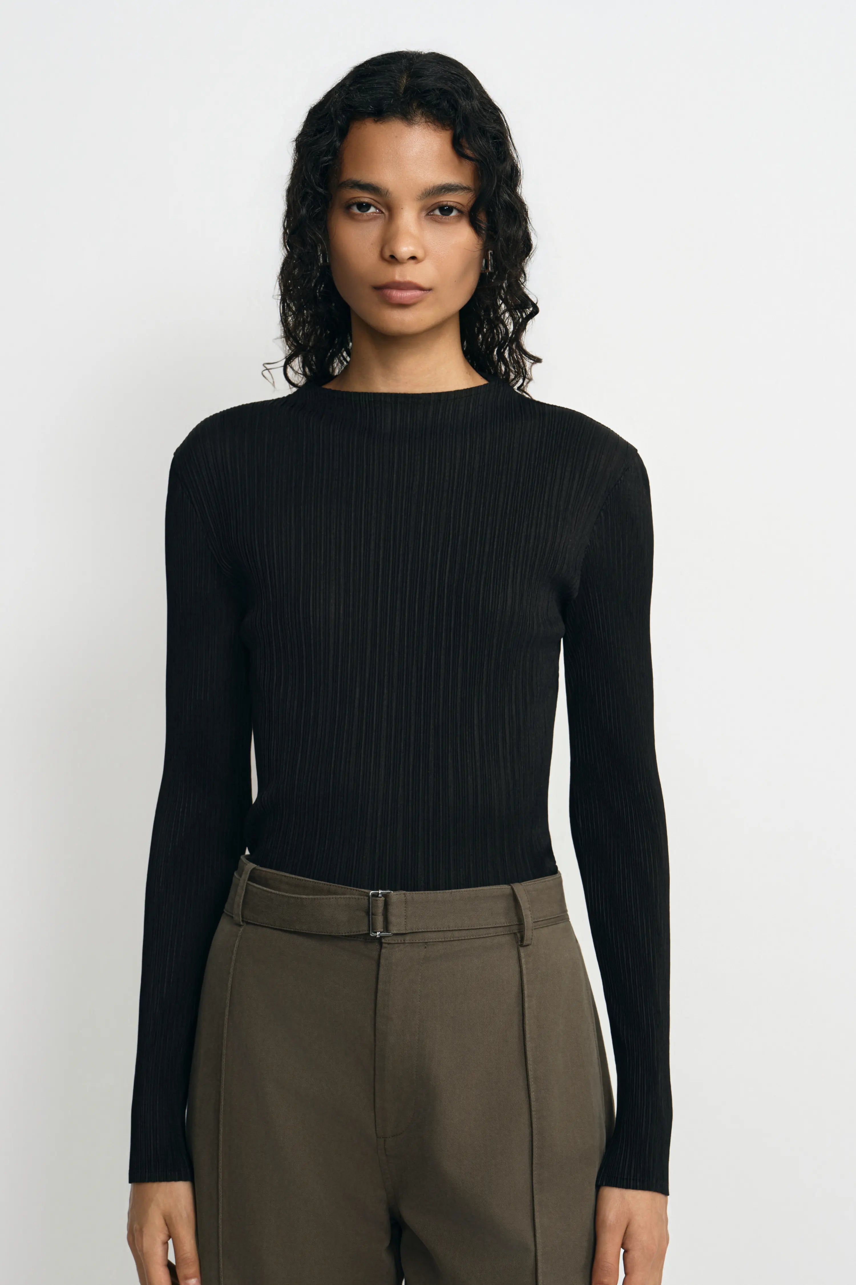 A woman modeling a black long-sleeve ribbed knit top with a boat neckline, paired with olive green trousers, against a plain light gray background.