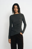 Front close-up of a model in a charcoal-heather long-sleeve top, showing the boat neckline, horizontal waist gathers and slanting hem over brown trousers.