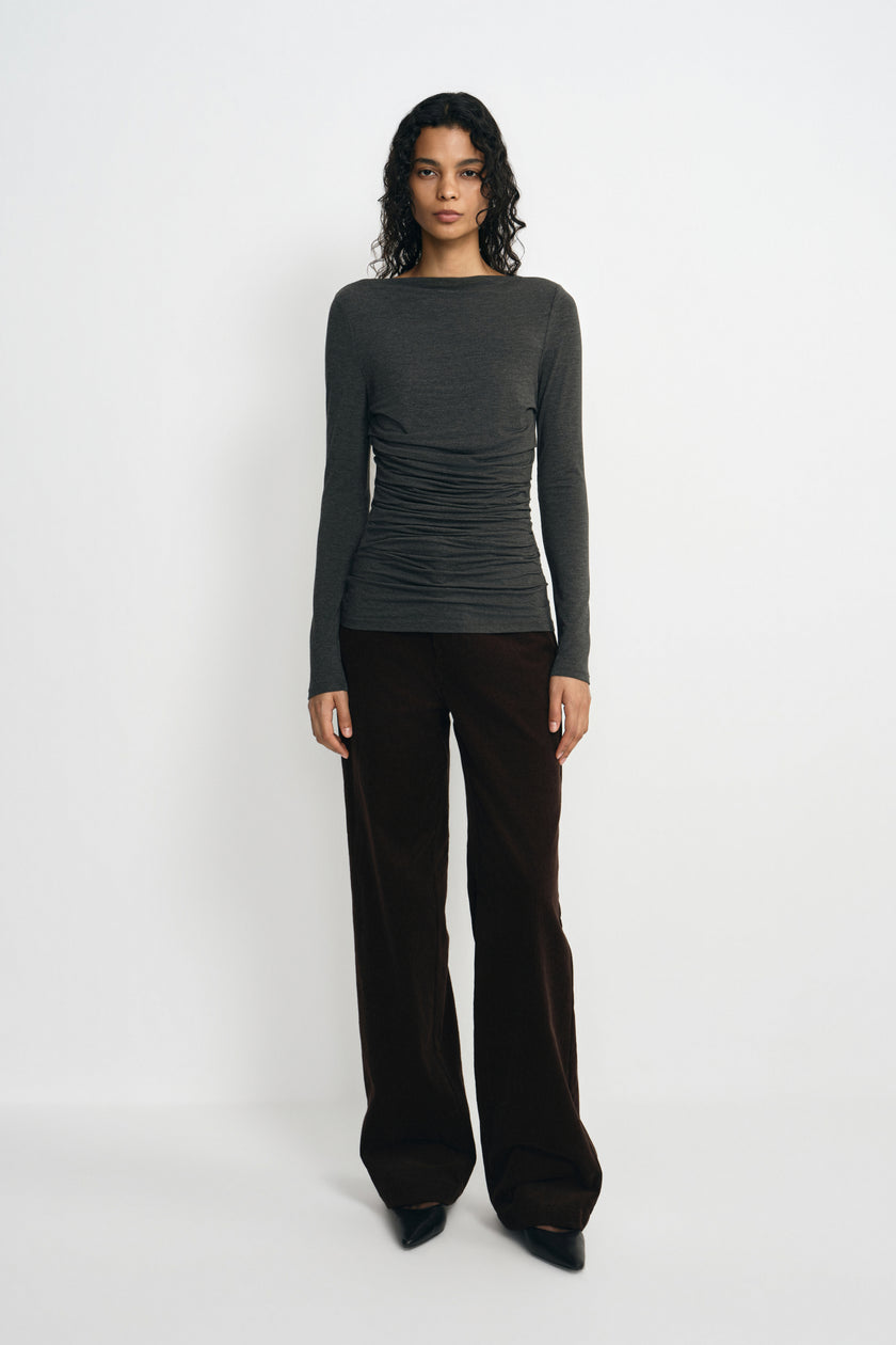 A model wearing a charcoal-heather boatneck top with a gathered waist and long sleeves, untucked over dark brown wide-leg trousers with black shoes.