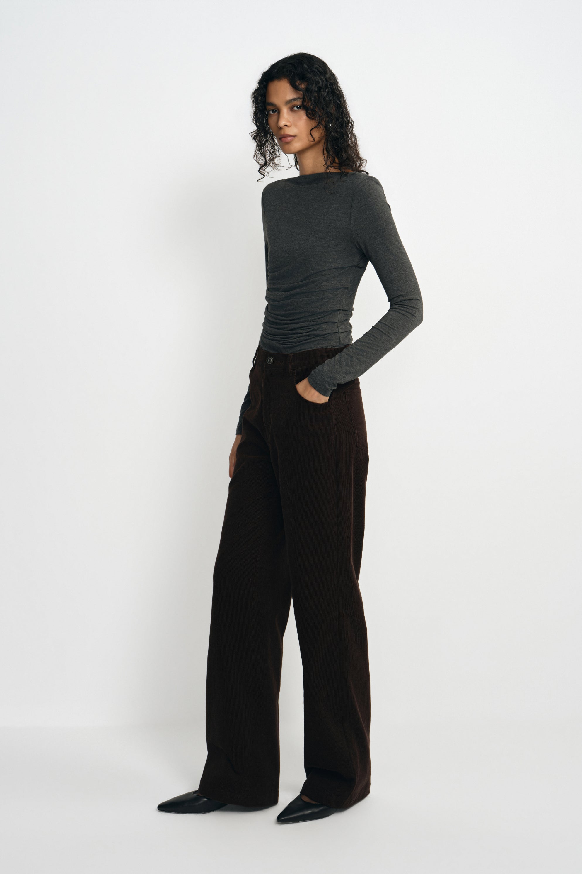 A model wearing a charcoal-heather long-sleeve top with a boat neck and gathered waist, tucked into dark brown wide-leg trousers with black pointed shoes.