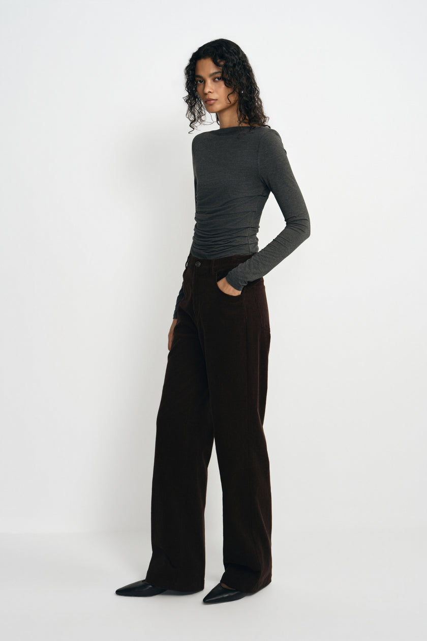 A model wearing a charcoal-heather long-sleeve top with a boat neck and gathered waist, tucked into dark brown wide-leg trousers with black pointed shoes.