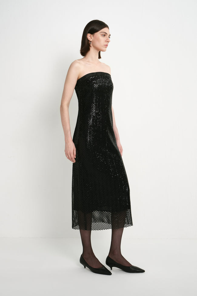 A model wearing the Rue Sophie Céleste Sequin Crochet Mesh Dress in black with a straight strapless neckline, fitted silhouette, and shimmering mesh finish, three-quarter view.
