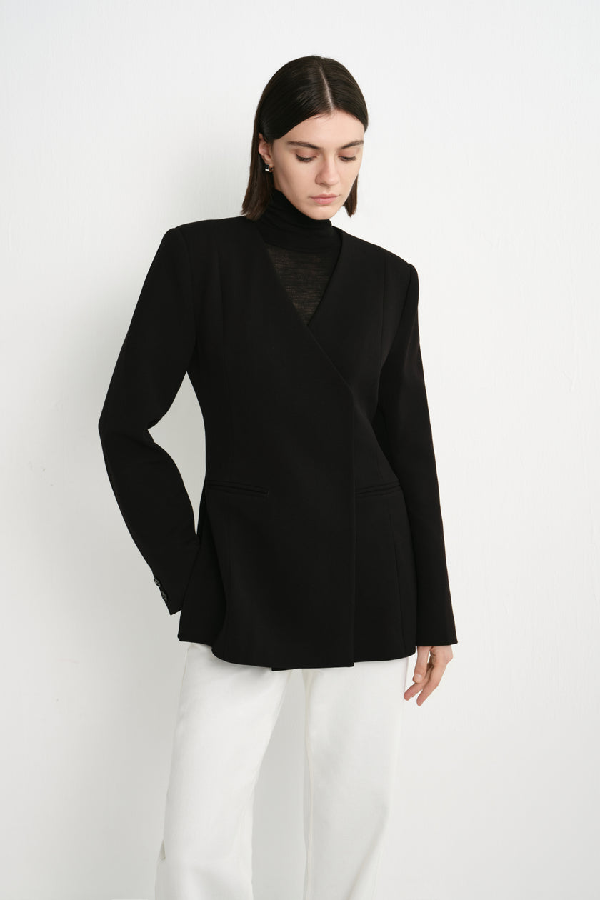 A model wearing the Rue Sophie Marion Side Slit Jacket in black, featuring a deep V-neckline, structured shoulders, welt pockets, and a defined waist, three-quarter front view.