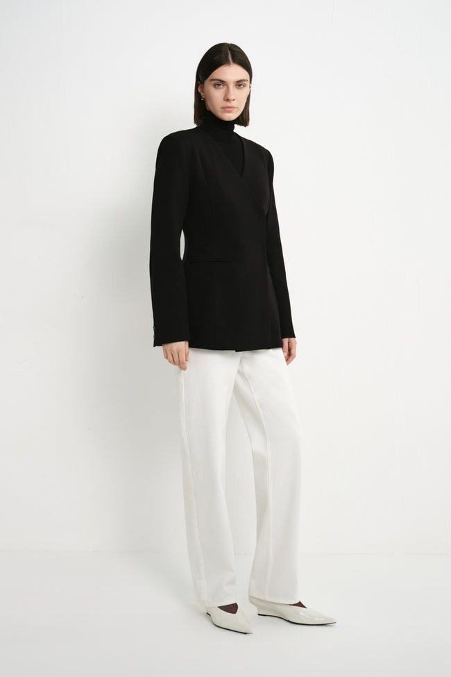 A model wearing the Rue Sophie Marion Side Slit Jacket in black with a collarless V-neckline, long sleeves, structured shoulders, and a tailored waist, three-quarter view.