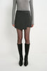 A model wearing the Rue Sophie Alina Asymmetric Mini Skirt in H. Charcoal with a structured wrap-style front panel and asymmetric hem, full-length front view.