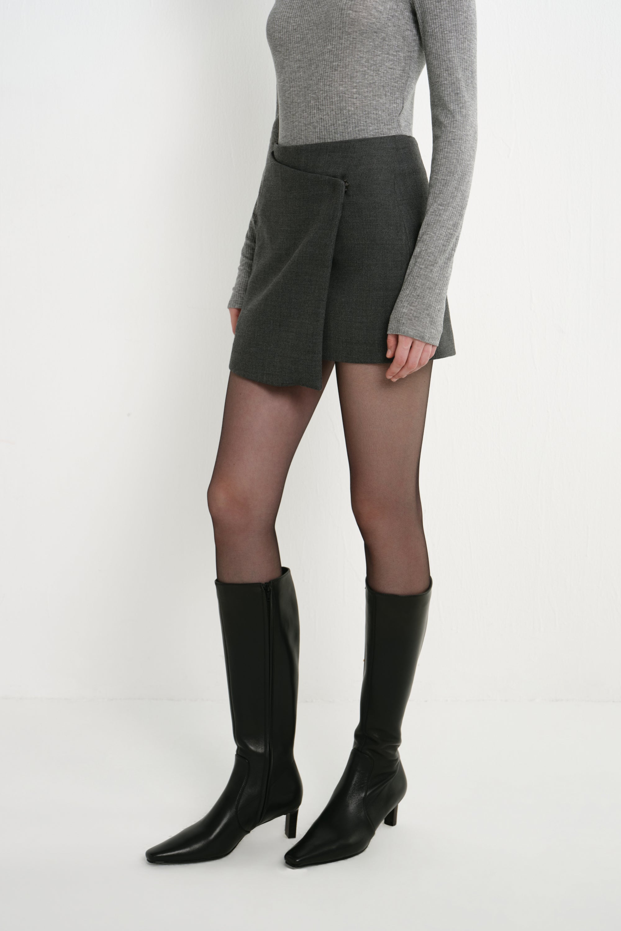A model wearing the Rue Sophie Alina Asymmetric Mini Skirt in H. Charcoal, highlighting the angled wrap-front panel and structured mini silhouette, three-quarter view.