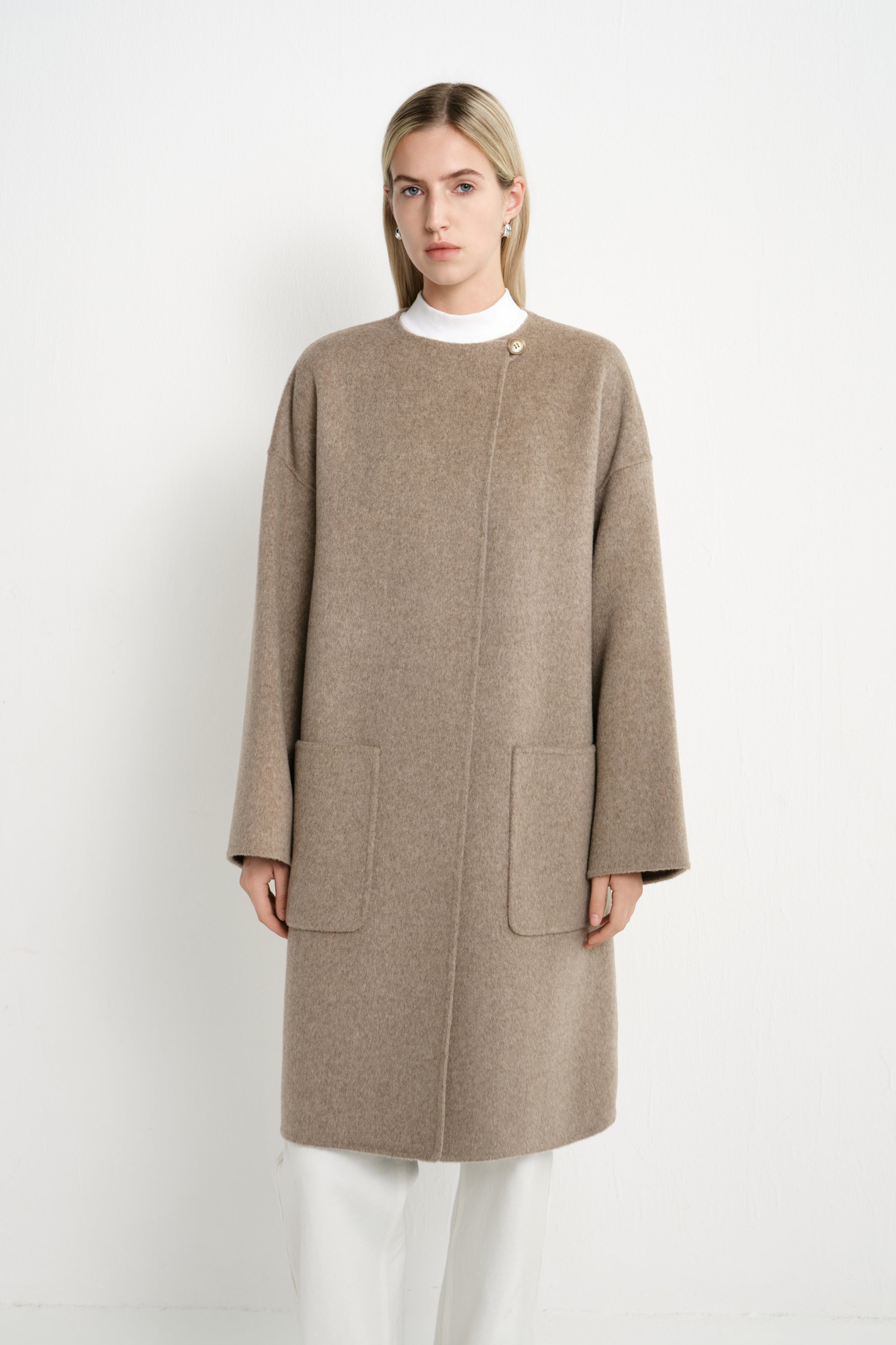 A model wearing a Rue Sophie oak collarless long coat with a single neck button, concealed front closure, and oversized patch pockets, front view.