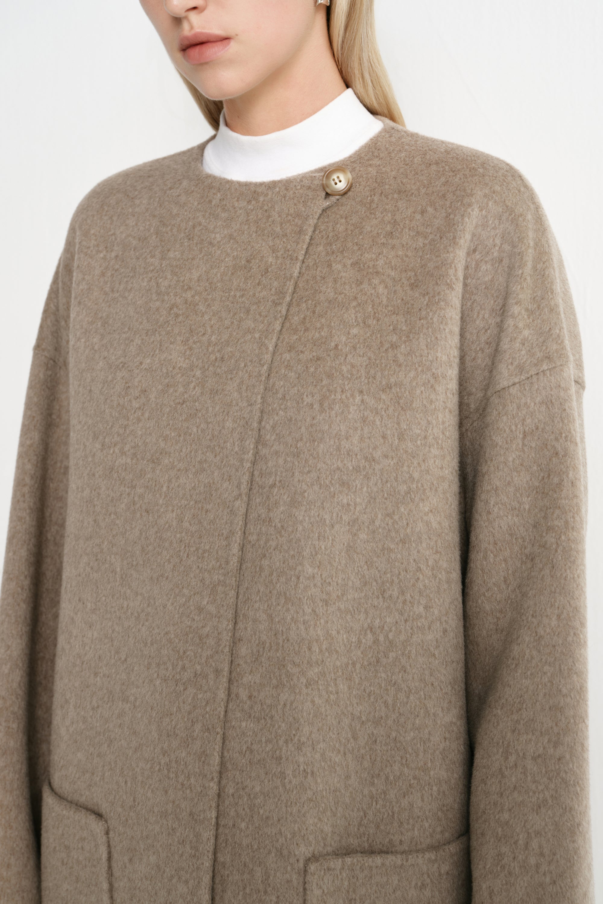 Close-up of a Rue Sophie oak collarless long coat featuring a single neck button, concealed front closure, drop shoulder construction, and soft brushed texture.