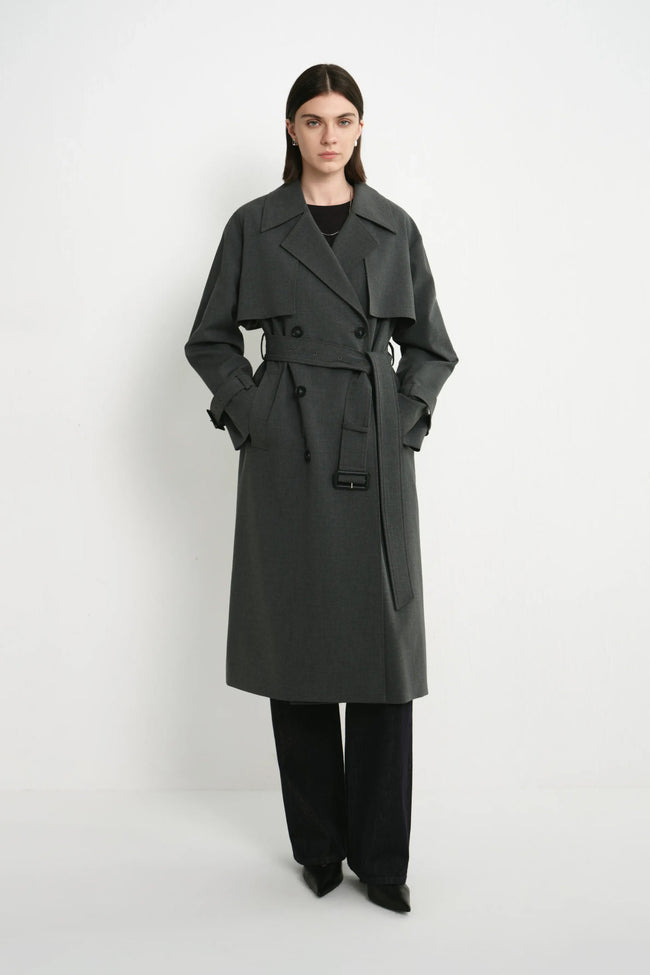 A model wearing the Rue Sophie Gavin Trench Coat in H. Charcoal with wide lapels, double-breasted buttons, a belted waist, and a relaxed tailored fit, three-quarter front view.