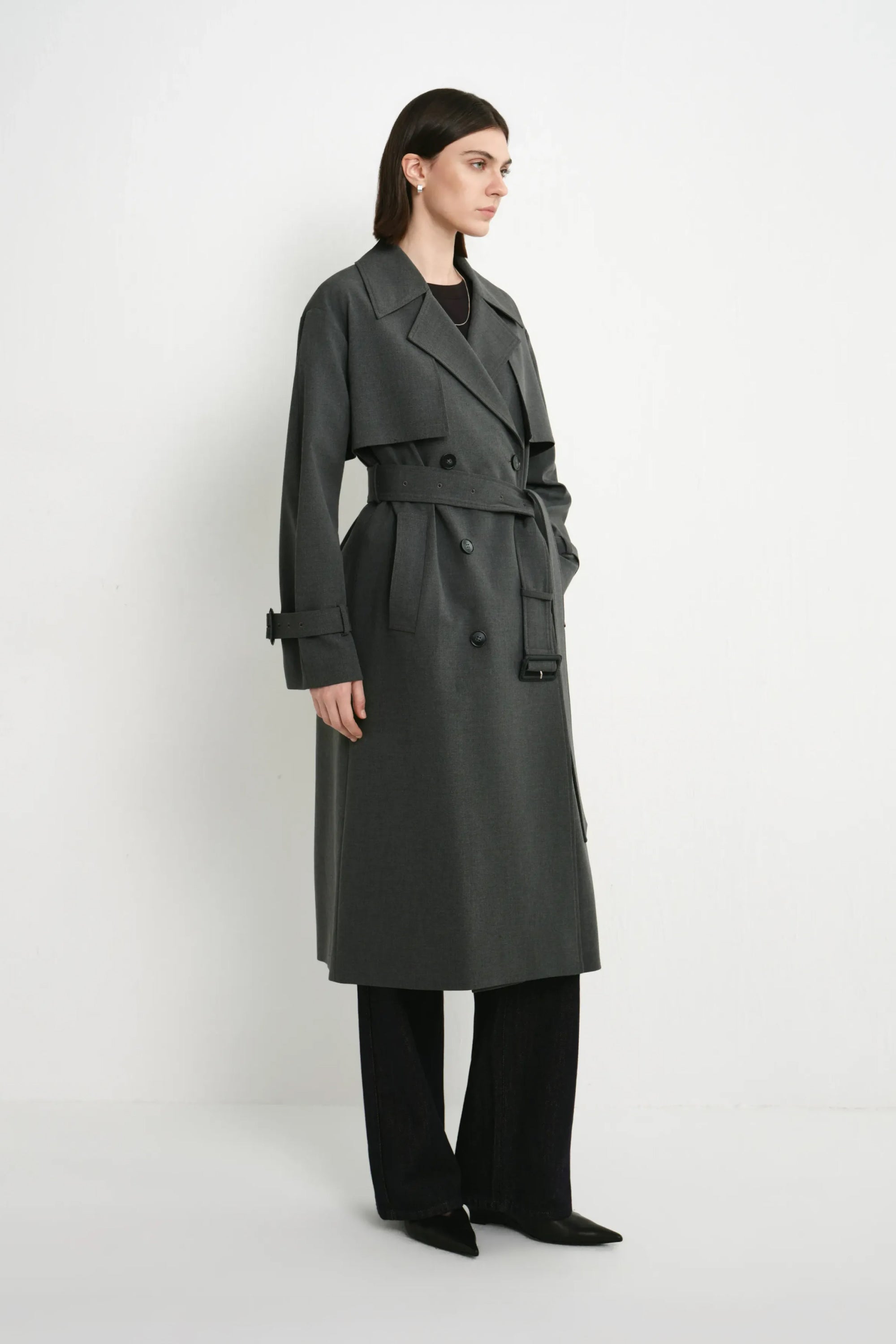 A model wearing the Rue Sophie Gavin Trench Coat in H. Charcoal, featuring a structured collar, double-breasted closure, waist belt, and long tailored silhouette, side view.