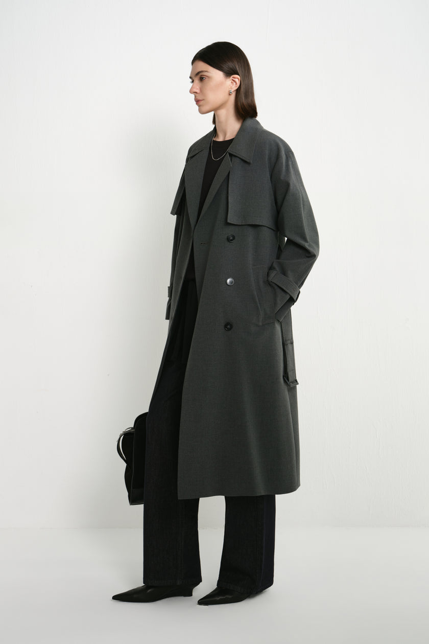 A model wearing the Rue Sophie Gavin Trench Coat in H. Charcoal, featuring wide lapels, a double-breasted front, belted waist, side pockets, and a relaxed long silhouette, three-quarter side view.