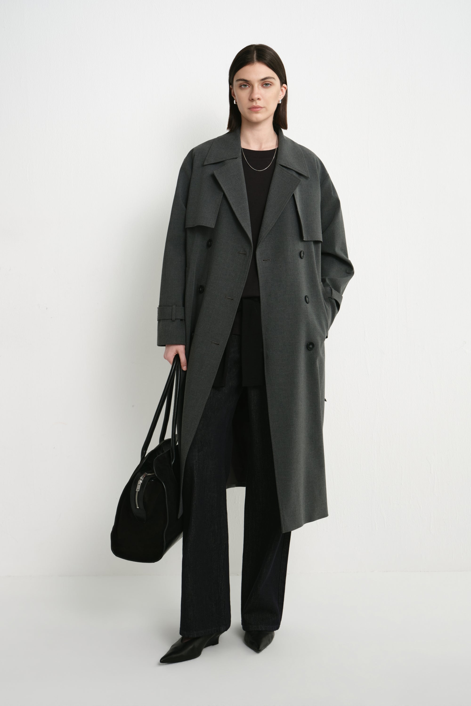 A model wearing the Rue Sophie Gavin Trench Coat in H. Charcoal, featuring a double-breasted front, wide lapels, belted waist, and a relaxed long silhouette, front view.