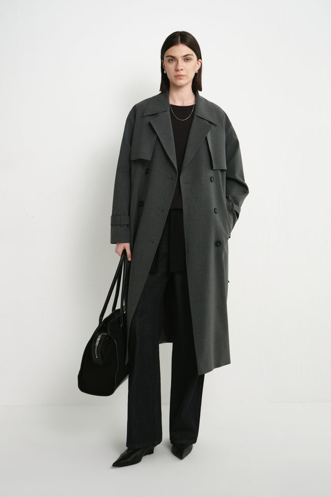 A model wearing the Rue Sophie Gavin Trench Coat in H. Charcoal, featuring a double-breasted front, wide lapels, belted waist, and a relaxed long silhouette, front view.