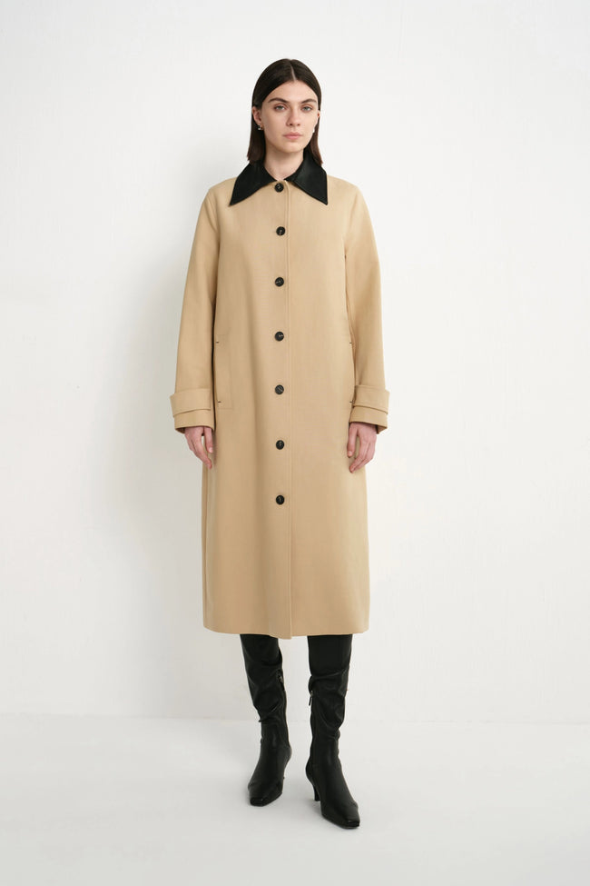 A woman stands against a plain white studio background wearing a long beige trench coat with a contrasting black collar and black buttons, paired with black knee-high boots.
