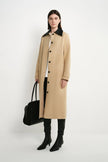 A model stands against a plain white studio background wearing a long beige coat with a black collar, black pants, black knee-high boots, and holding a black handbag.