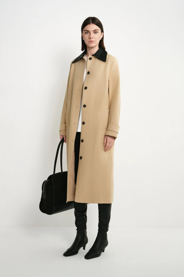 A model stands against a plain white studio background wearing a long beige coat with a black collar, black pants, black knee-high boots, and holding a black handbag.