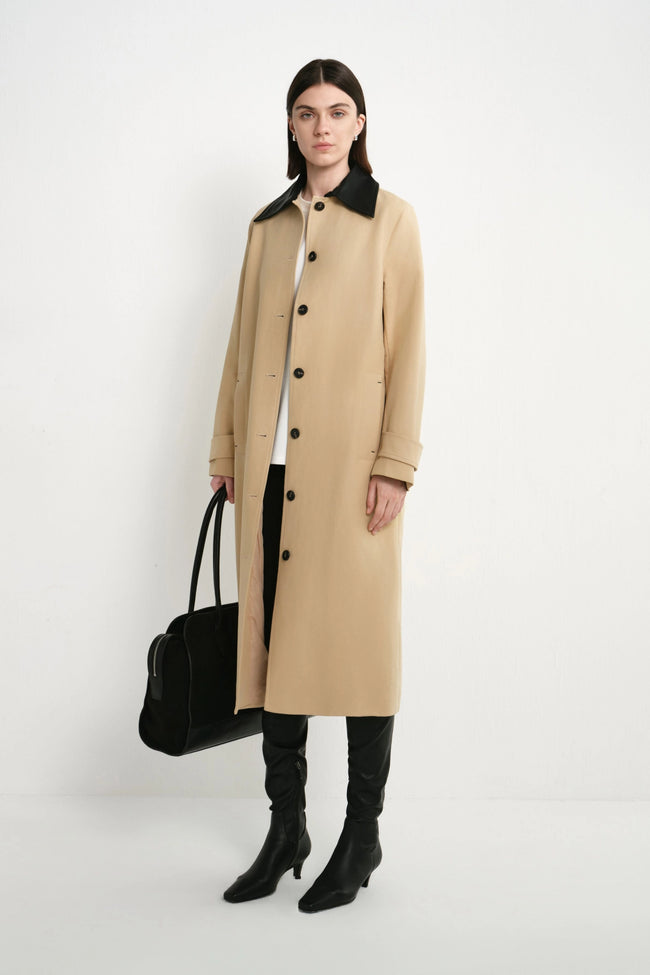 A model stands against a plain white studio background wearing a long beige coat with a black collar, black pants, black knee-high boots, and holding a black handbag.