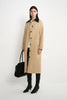 A model stands against a plain white studio background wearing a long beige coat with a black collar, black pants, black knee-high boots, and holding a black handbag.