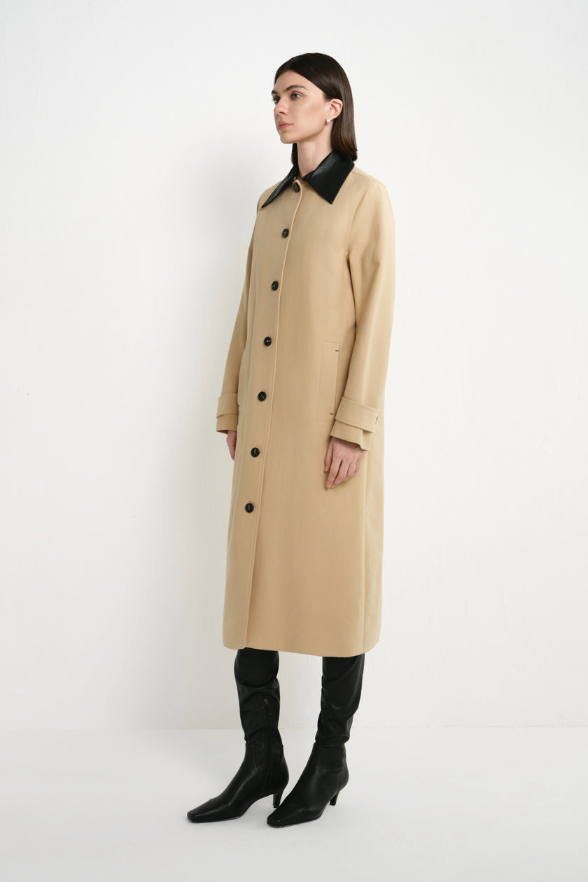 A woman modeling a long camel-colored trench coat with a contrasting black collar, black buttons, and black knee-high boots, standing against a plain white studio background.