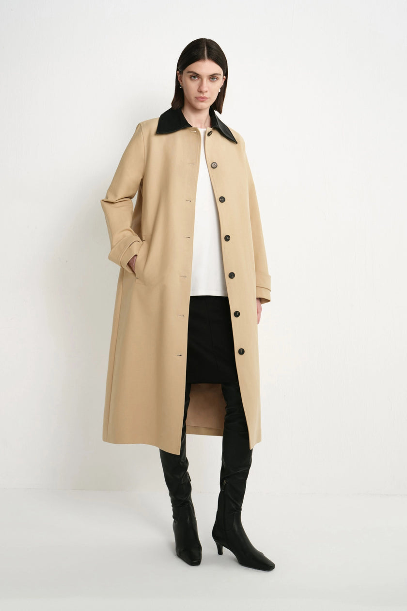A model stands against a plain white studio background wearing a long beige trench coat with a black collar, a white top, black pants, and black knee-high heeled boots.