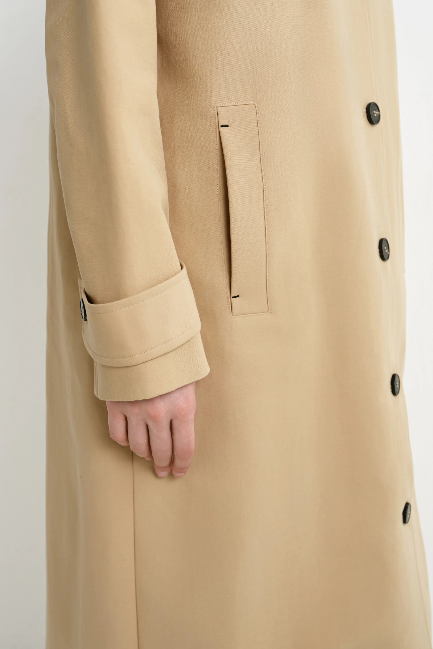 Close-up of a beige trench coat worn by a model, showing the sleeve with a button cuff, a vertical welt pocket, and dark buttons on the front closure.