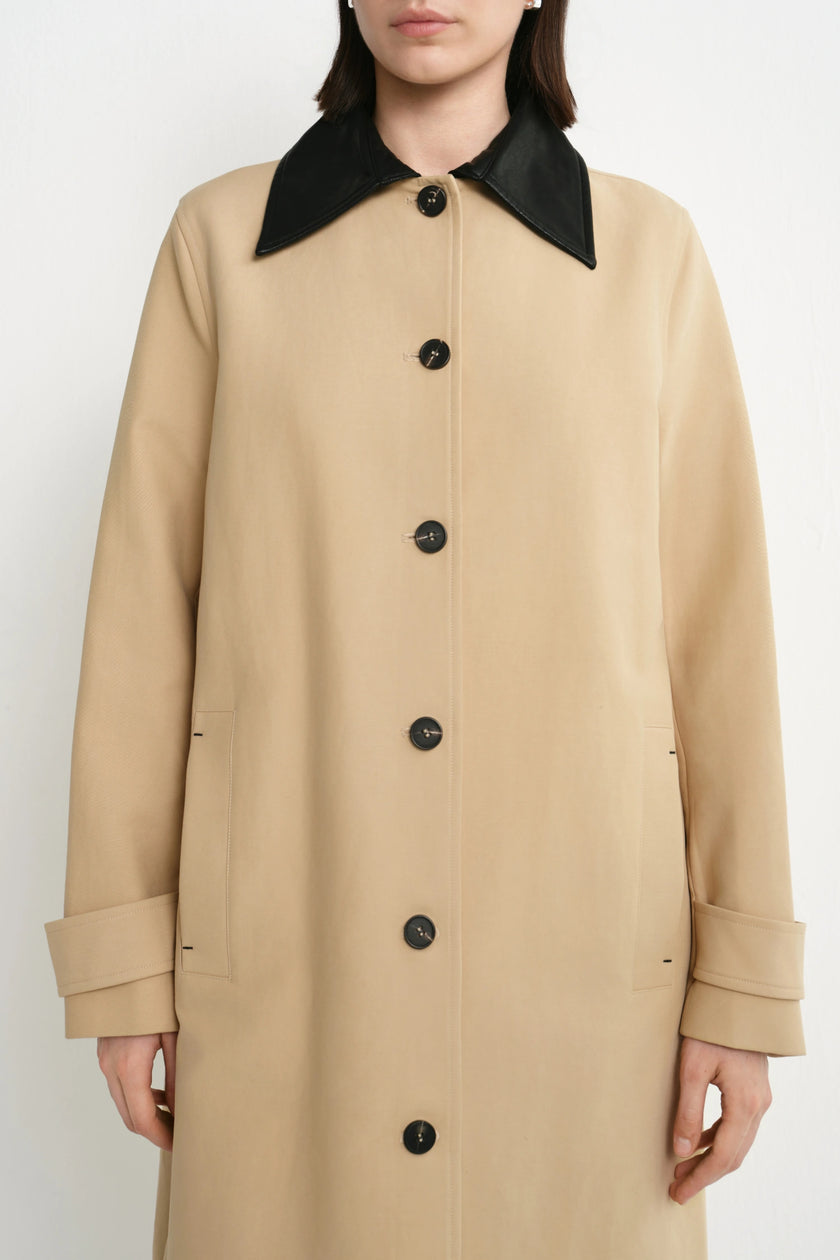 A model wearing a beige coat with a contrasting black collar and black buttons, shown from the neck down against a plain light background.