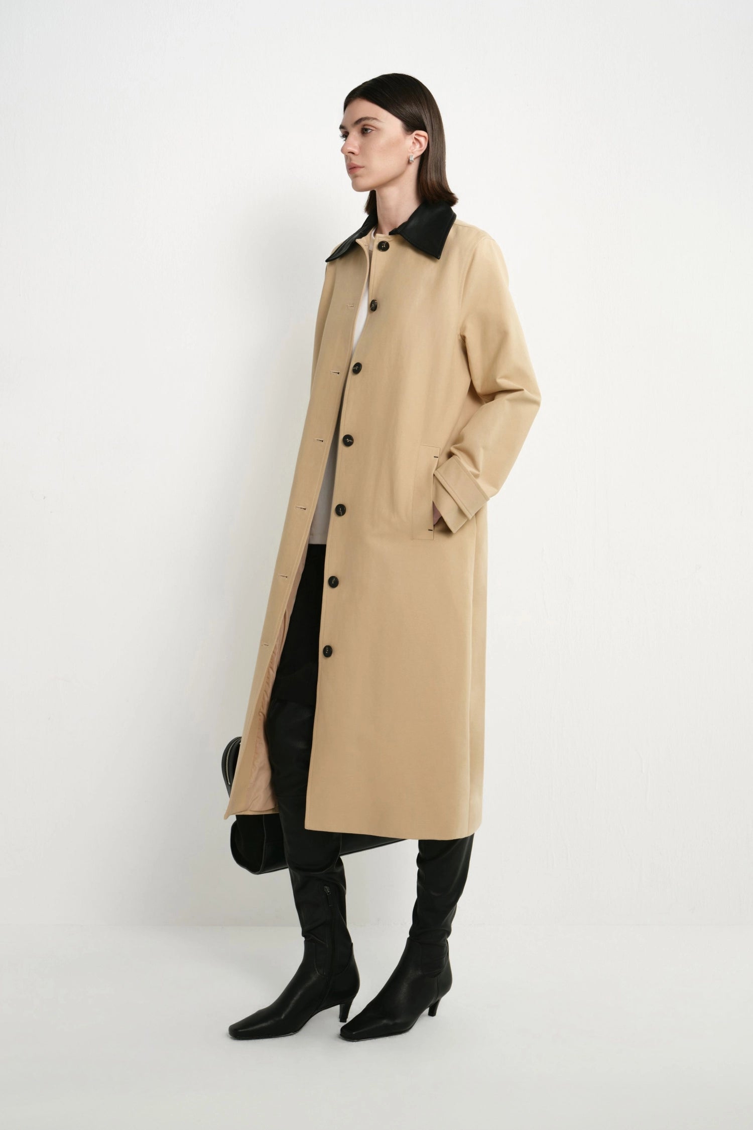 A woman models a long beige trench coat with a black collar, black buttons, and a black interior lining, styled with black pants and black knee-high boots against a plain white studio background.