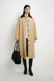 A model stands against a plain white studio background wearing a long beige trench coat with a contrasting black collar, black pants, black knee-high boots, and holding a black handbag.
