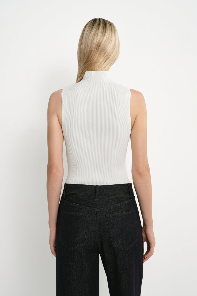 A woman shown from the back wearing a sleeveless white mock-neck top and dark charcoal pants against a plain white studio background.